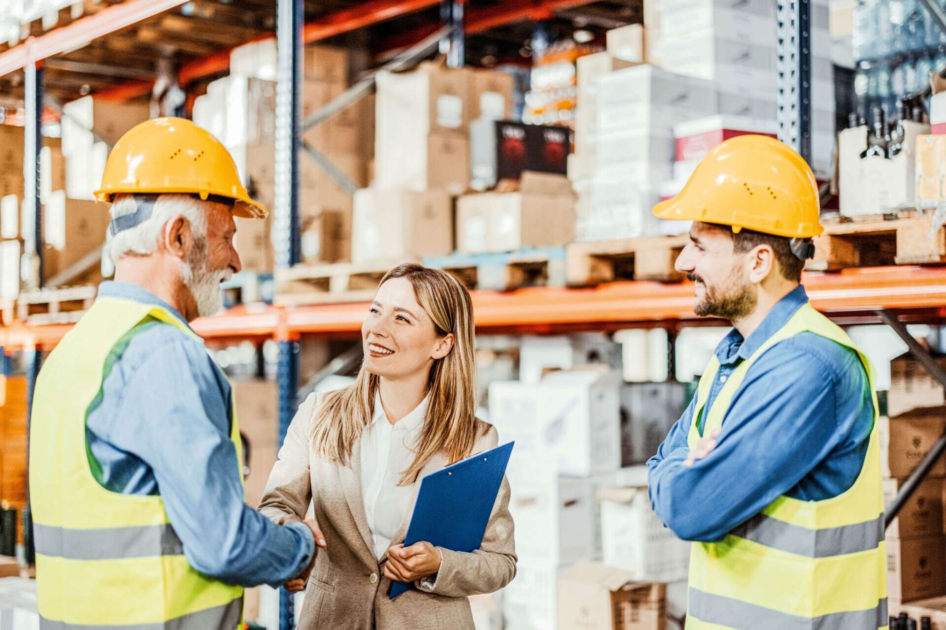 Drei Personen in einem Lager. Die Leiterin schüttelt dem älteren Mitarbeiter die Hand.
