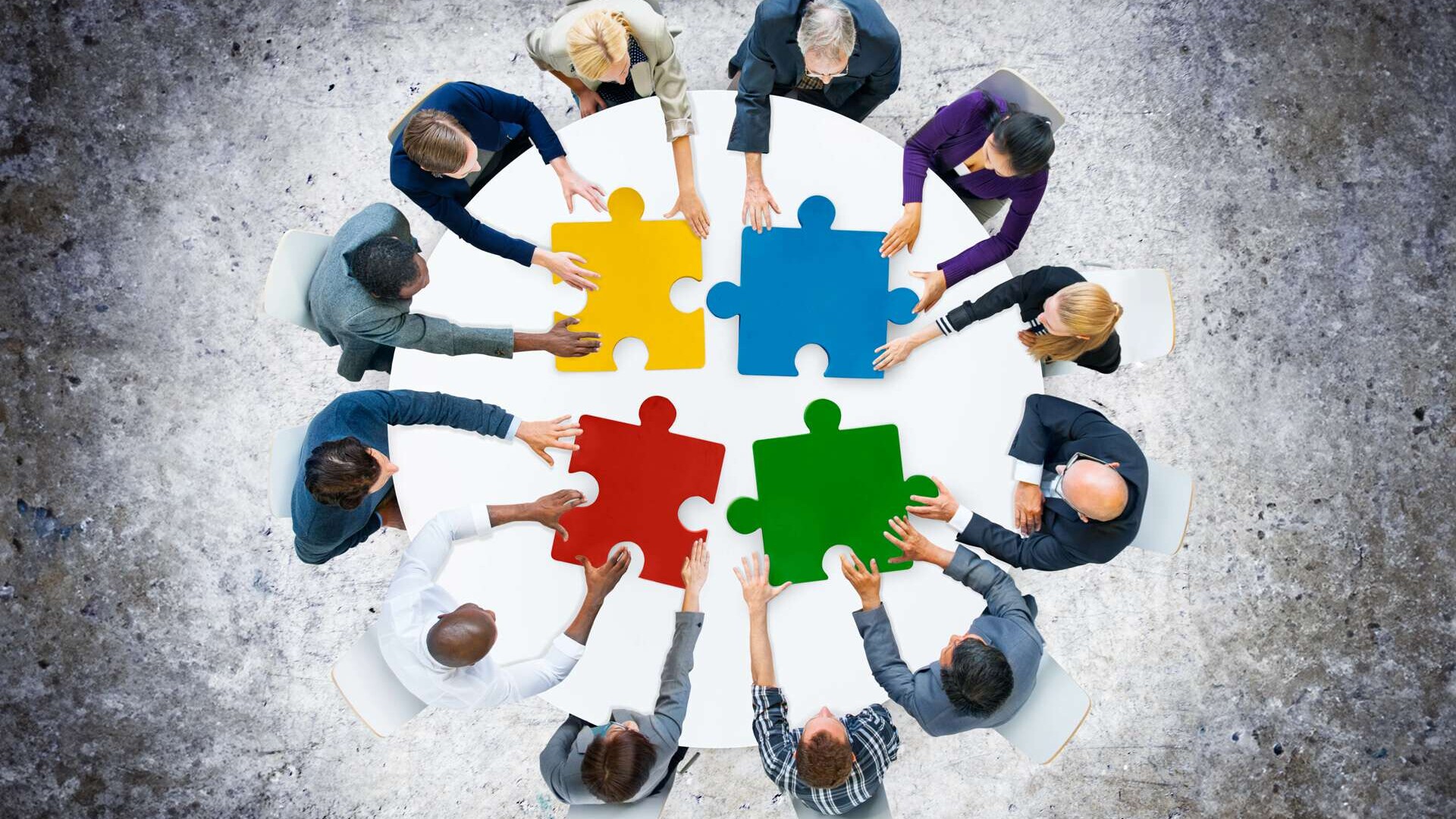 Symbolbild: mehrere Personen an einem runden Tisch, aus Vogelperspektive gesehen. Die Personen halten ihre Hände an überdimensionierten, bunten Puzzle-Teilen, die auf dem Tischblatt liegen.  #filzer