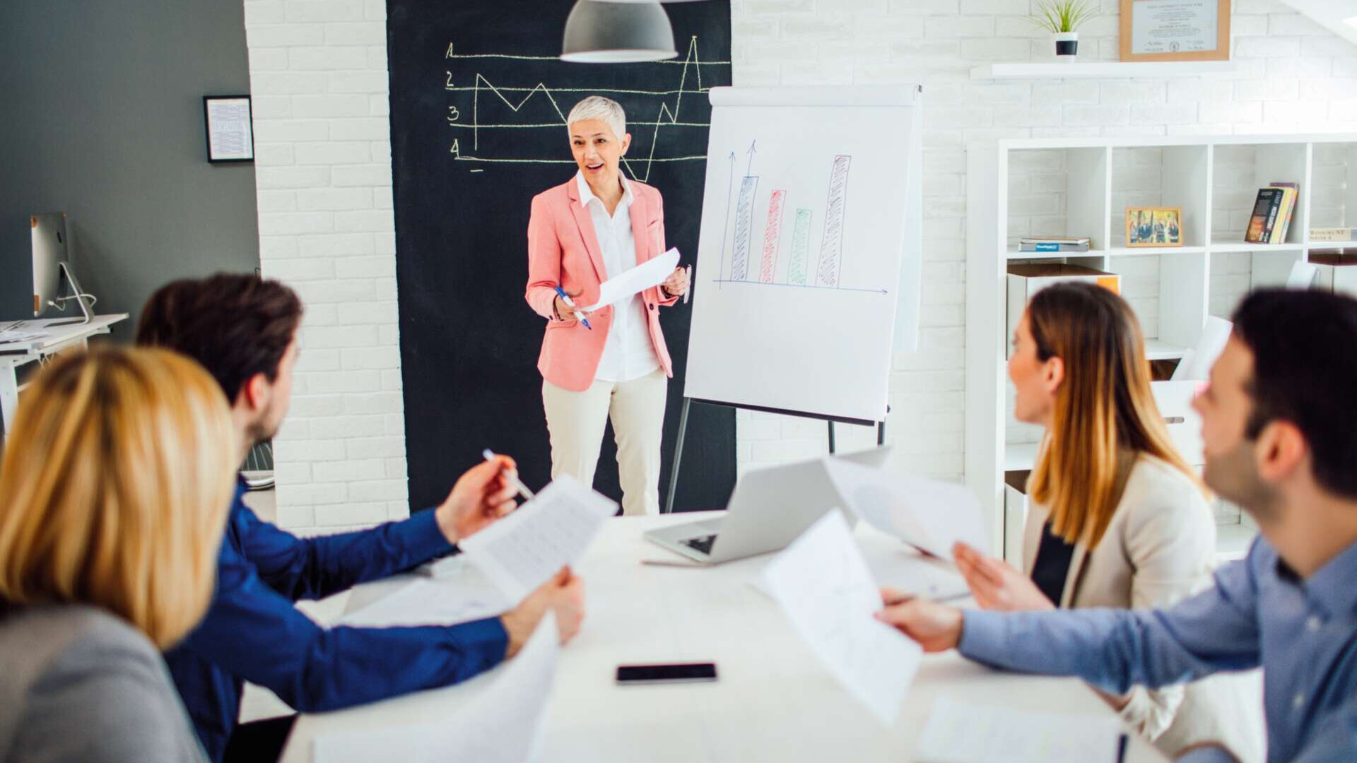Besprechung - eine Frau steht am Flipchart und hält einen Vortrag und eine Frau und zwei Männer sitzen am Tisch
