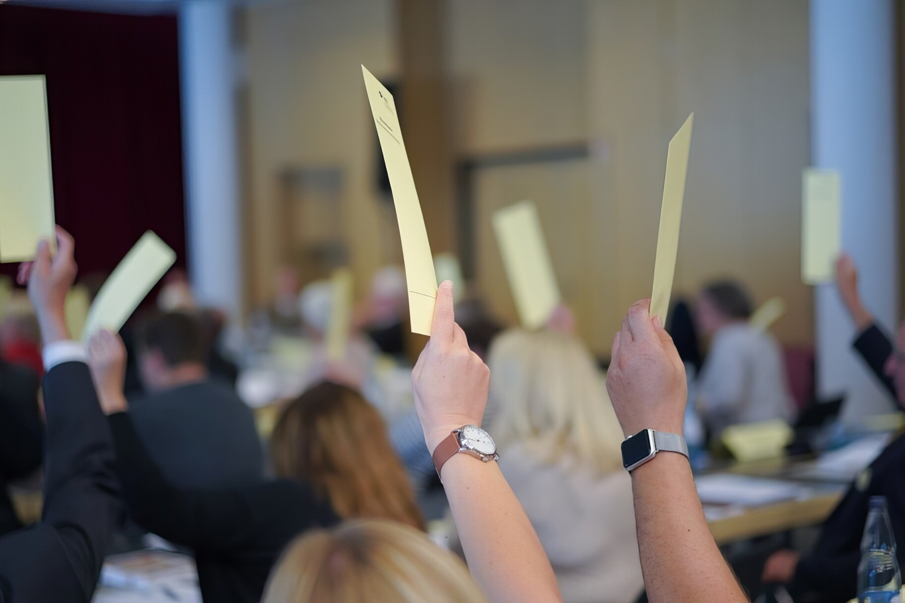 Mehrere sitzende Personen in einem Raum heben Zettel zur Abstimmung hoch.