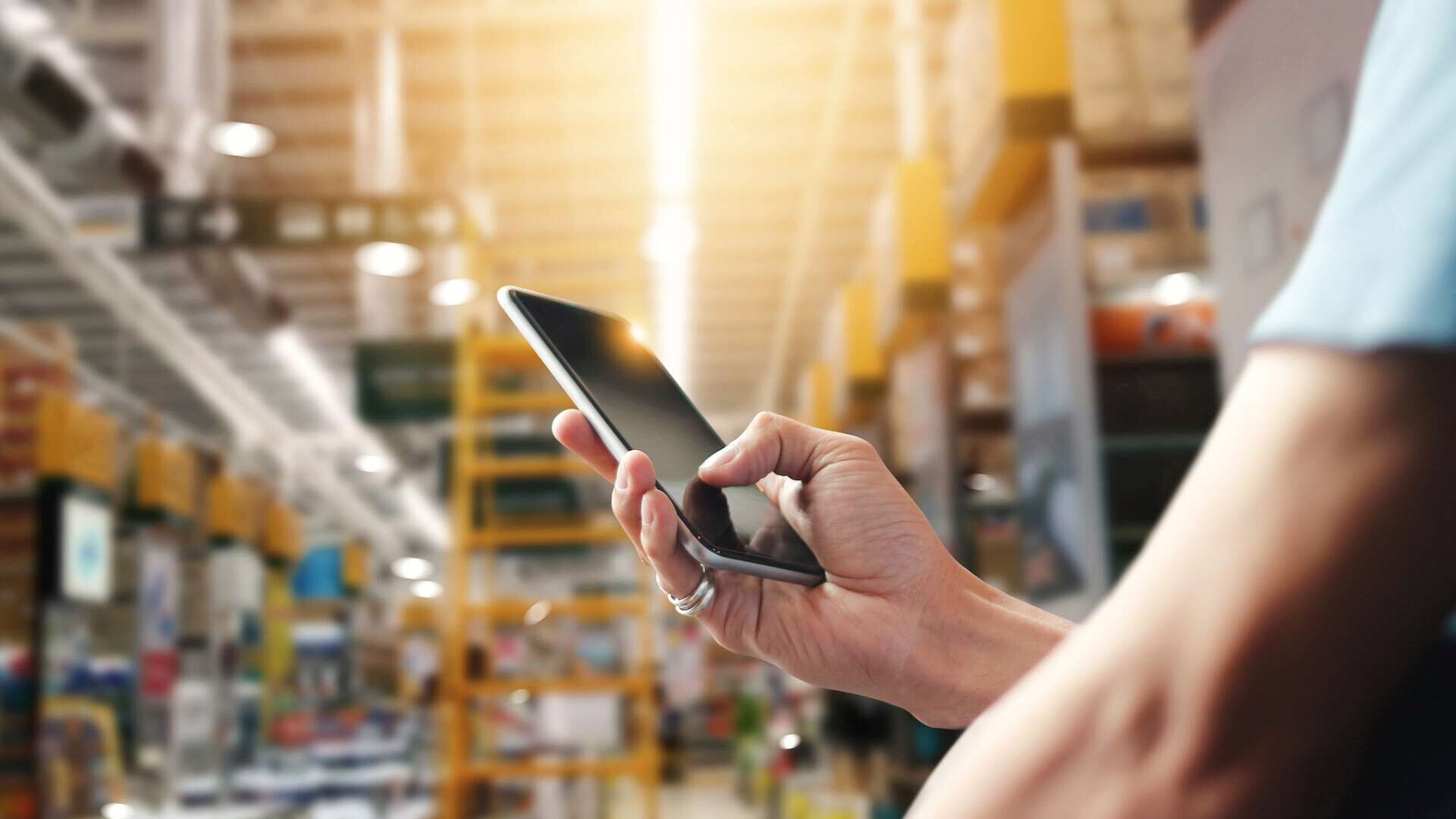 Großaufnahme einer Hand, die ein Smartphone hält; die Person selbst ist durch den Bildrand abgeblendet; im Hintergrund, im leichten Unschärfebereich, eine Fabrik- oder Lagerhalle. Originalbeschreibung iStock: "Factory worker using application on mobile smartphone to operate automation for modern trade. Checking order in large warehouse. Import and export the shipping cargo. Model Released (MR)."